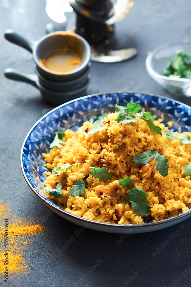 Indian Pilaf with Cauliflower yellow coloured in a blue plate on a gray table. 45 angle view. There are bowls of herbs and sprinkled seasoning on the table Indian Pilaf with Cauliflower yellow coloured in a blue plate on a gray table. 45 angle view. There are bowls of herbs and sprinkled seasoning on the table