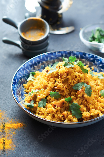 Indian Pilaf with Cauliflower yellow coloured in a blue plate on a gray table. 45 angle view. There are bowls of herbs and sprinkled seasoning on the table