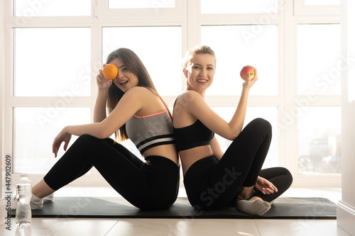 Sportswomen sit on fitness mat and hold fruits