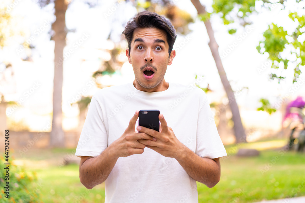 Young caucasian man in a park looking at the camera while using the mobile with surprised expression Young caucasian man in a park looking at the camera while using the mobile with surprised expression