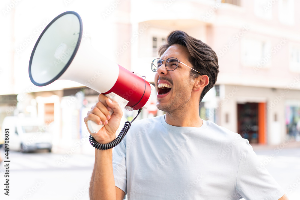 Young caucasian man at outdoors shouting through a megaphone to announce something in lateral position Young caucasian man at outdoors shouting through a megaphone to announce something in lateral position