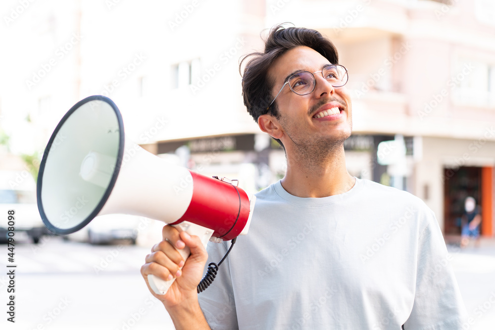 Young caucasian man at outdoors holding a megaphone and looking up while smiling Young caucasian man at outdoors holding a megaphone and looking up while smiling