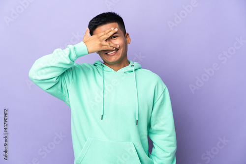 Young Ecuadorian man isolated on purple background covering eyes by hands and smiling © luismolinero