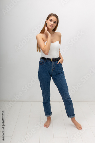 young attractive caucasian woman with long brown hair in corset, blue jeans on white studio background. skinny pretty lady posing with bare feet at bright room. portrait of beautiful female