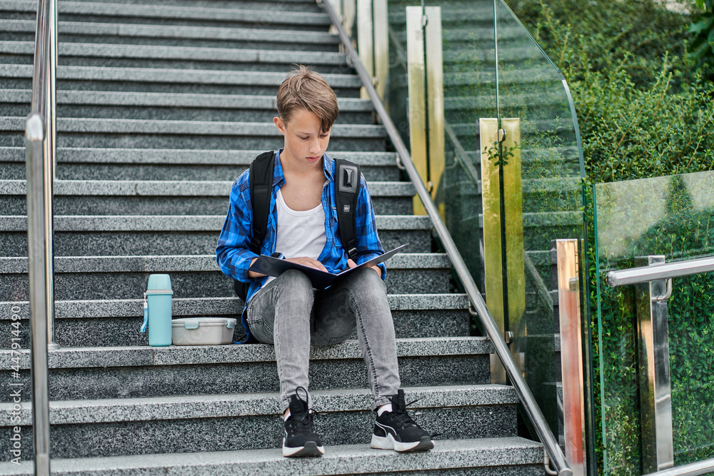 Little boy sitting on stairs near school and read book