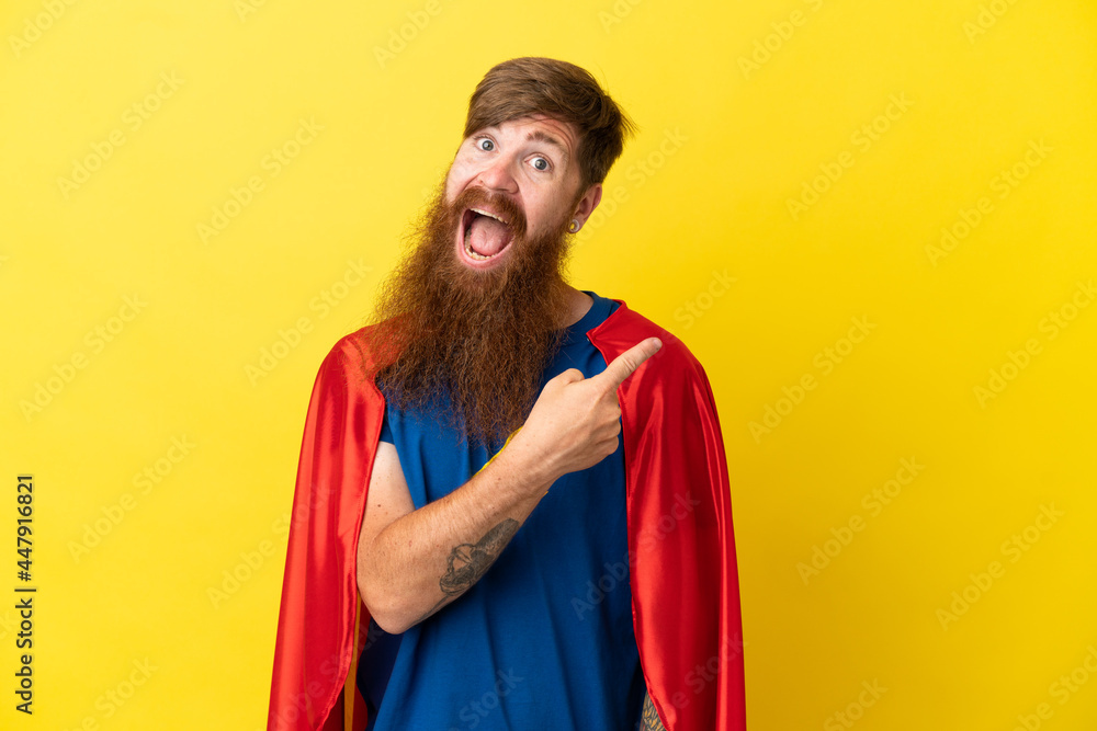 Redhead Super Hero man isolated on yellow background surprised and pointing side