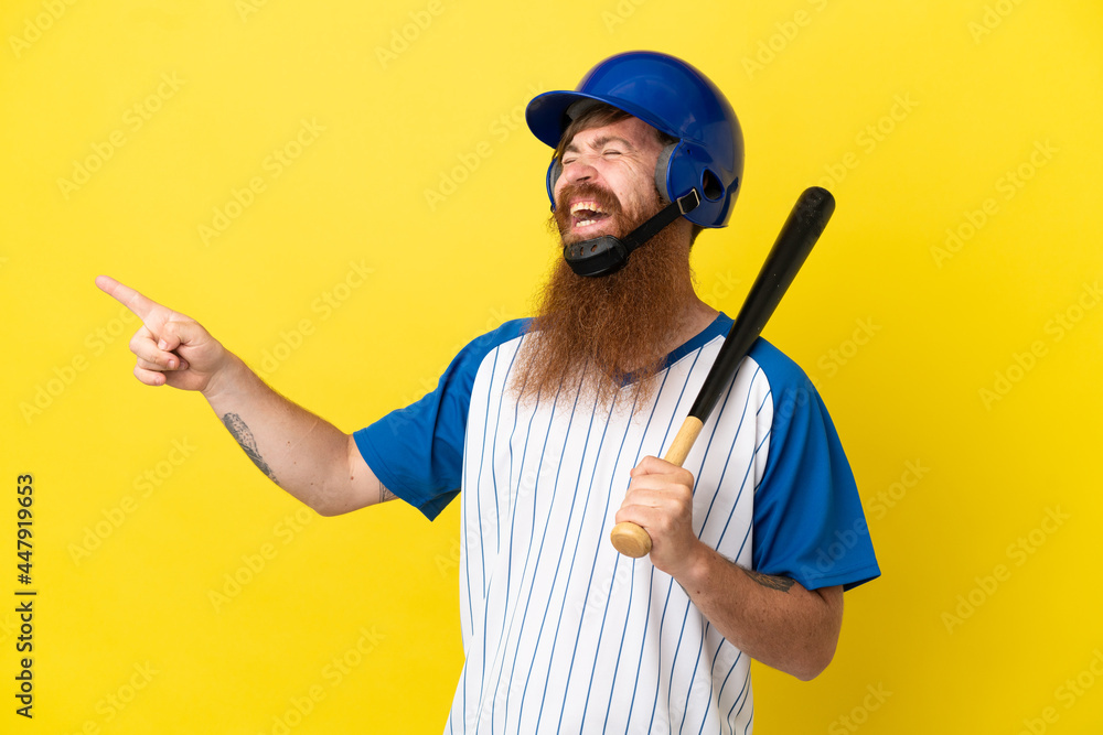 Redhead baseball player man with helmet and bat isolated on yellow background pointing finger to the side and presenting a product Redhead baseball player man with helmet and bat isolated on yellow background pointing finger to the side and presenting a product