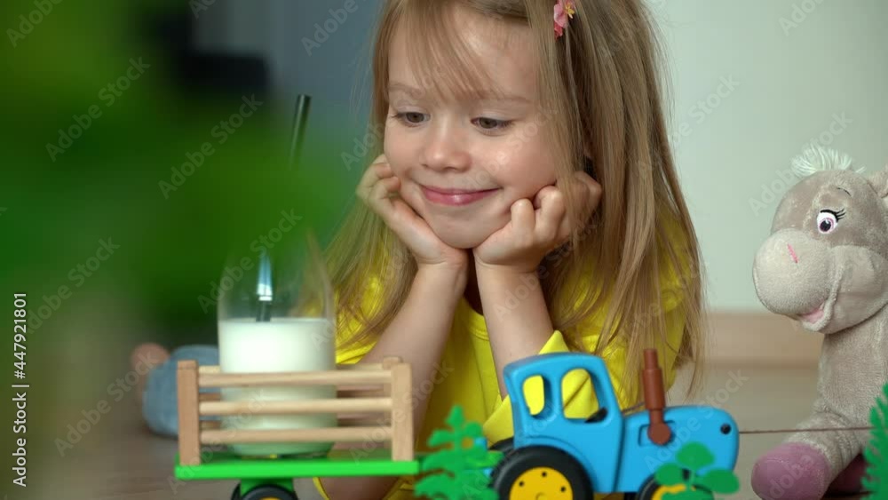 Kid plays in farm, toy tractor delivers milk in cart. Close up of little happy girl smiles and drinks milk from bottle. Food and drink concept Kid plays in farm, toy tractor delivers milk in cart. Close up of little happy girl smiles and drinks milk from bottle. Food and drink concept