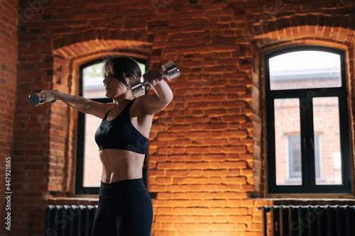 Shoot from below of young athletic woman with beautiful strong body wearing activewear holding dumbbells on retracted arms. Caucasian fitness female workout out exercising in dark gym.