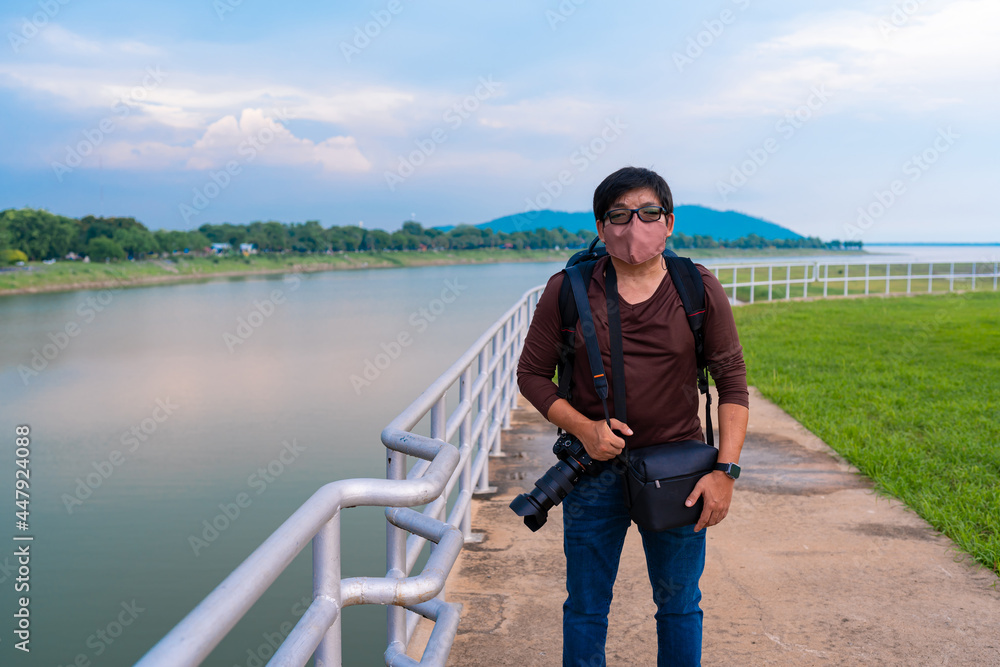 backpack Asian man with camera wear fabric protective face mask during travel at Pasak dam lake in Lopburi, Thailand. man relax on vacation standing alone 