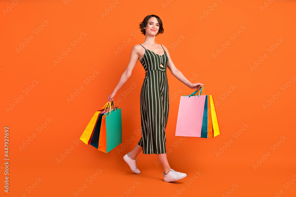 Full length photo of nice brunette millennial lady hold bags go wear overall sneakers isolated on orange background Full length photo of nice brunette millennial lady hold bags go wear overall sneakers isolated on orange background