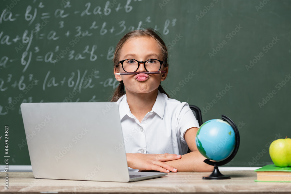 playful schoolgirl with pencil between lips and nose sitting near laptop, globe and blurred chalkboard playful schoolgirl with pencil between lips and nose sitting near laptop, globe and blurred chalkboard