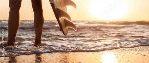 Close-up bottom pov view young adult female surfer girl legs with surfboard stand at ocean coast wave against warm sunrise or sunset sun. Sport healthy carefree freedom lifestyle vacation concept