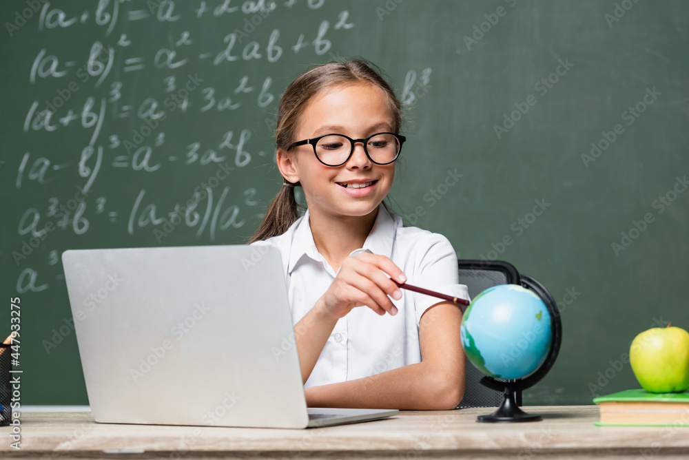 smiling schoolkid pointing with pencil at globe near laptop and blurred chalkboard smiling schoolkid pointing with pencil at globe near laptop and blurred chalkboard