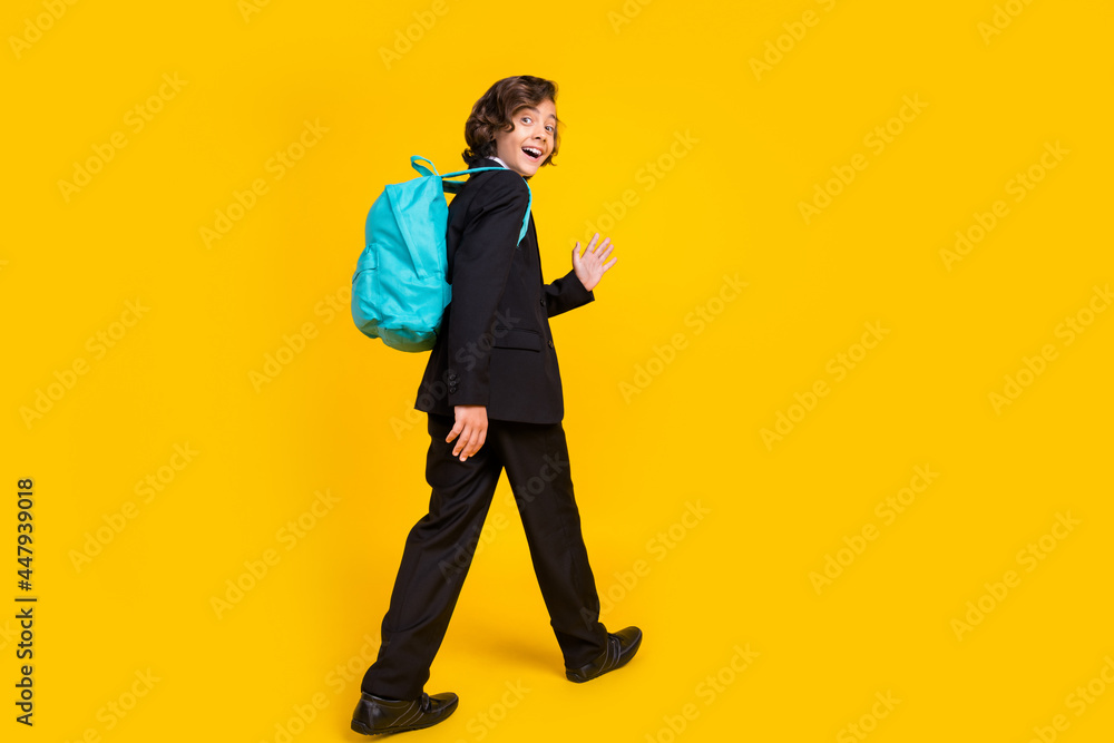 Photo of friendly little schoolboy walk wave palm wear backpack black uniform shoes isolated yellow color background Photo of friendly little schoolboy walk wave palm wear backpack black uniform shoes isolated yellow color background