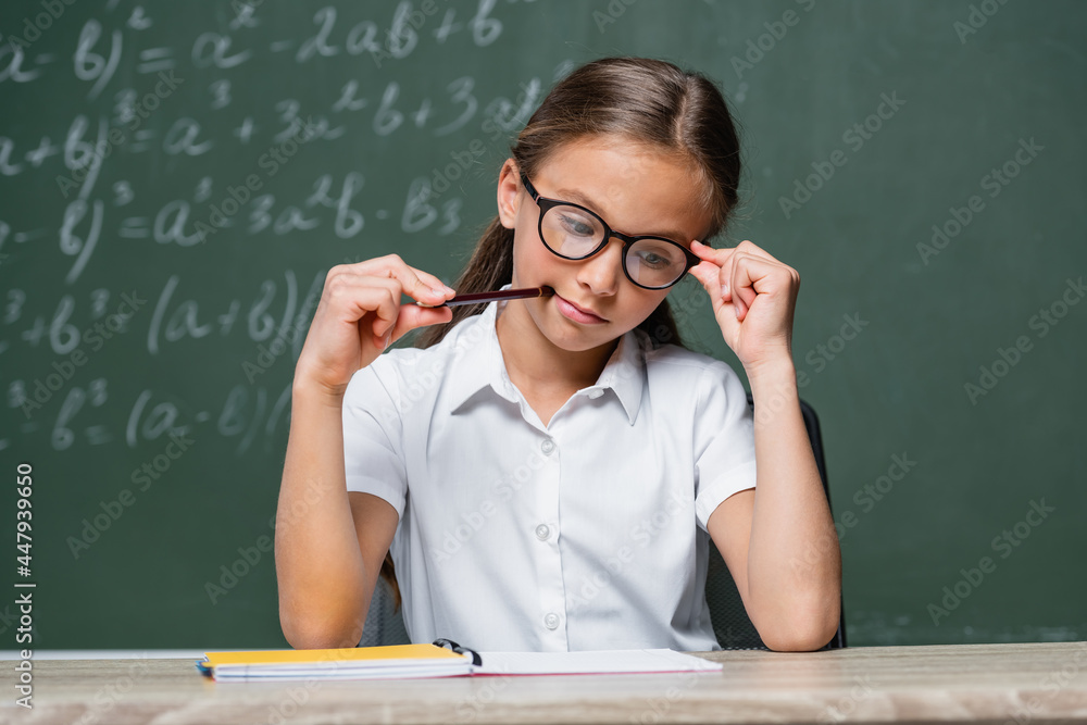 pensive schoolgirl holding pencil while thinking near notebook in classroom