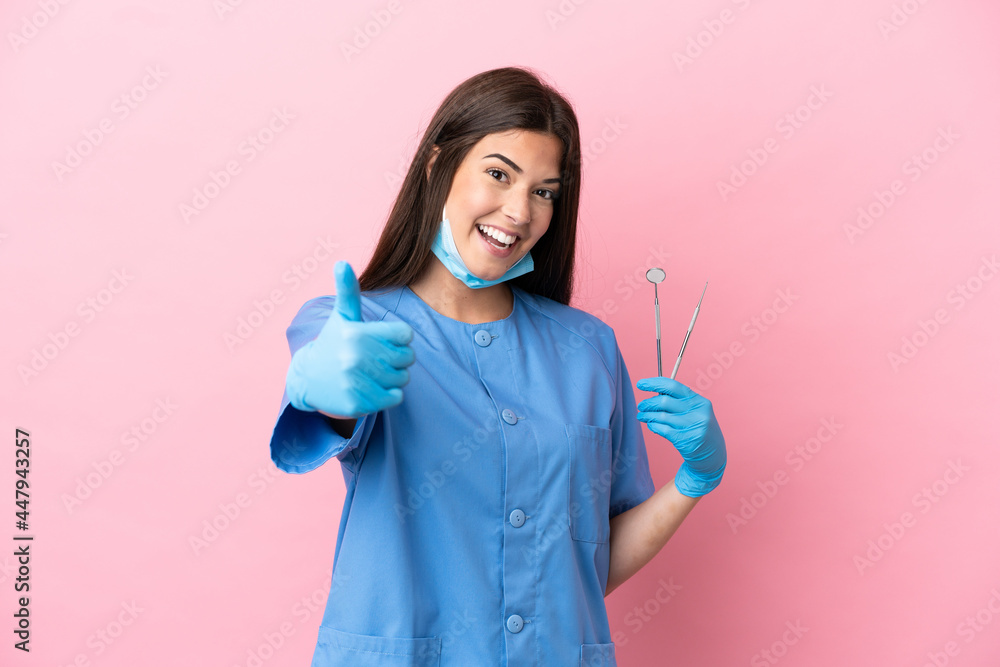 Dentist woman holding tools isolated on pink background with thumbs up because something good has happened Dentist woman holding tools isolated on pink background with thumbs up because something good has happened