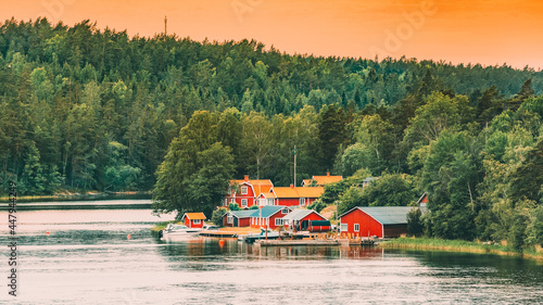 Sweden. Many Beautiful Red Swedish Wooden Log Cabins Houses On Rocky Island Coast In Summer Evening. Lake Or River Landscape