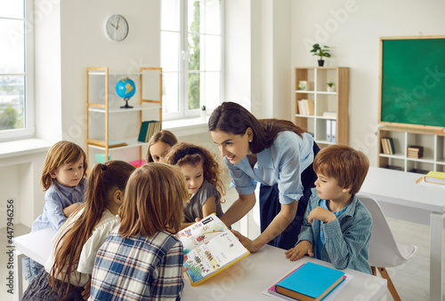 Young friendly caucasian female teacher in the classroom with a book in her hands teaches young students. Modern elementary school students stand around a desk and listen intently to the teacher.