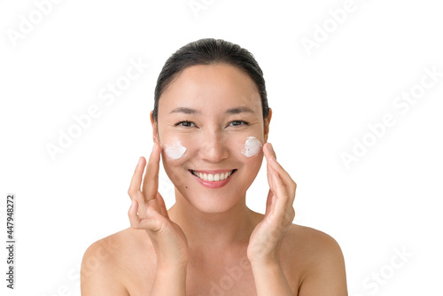 a young attractive Asian woman smiles and applies a white cream on her face on a white background. the concept of skin care, skin rejuvenation, preservation of youth and elasticity