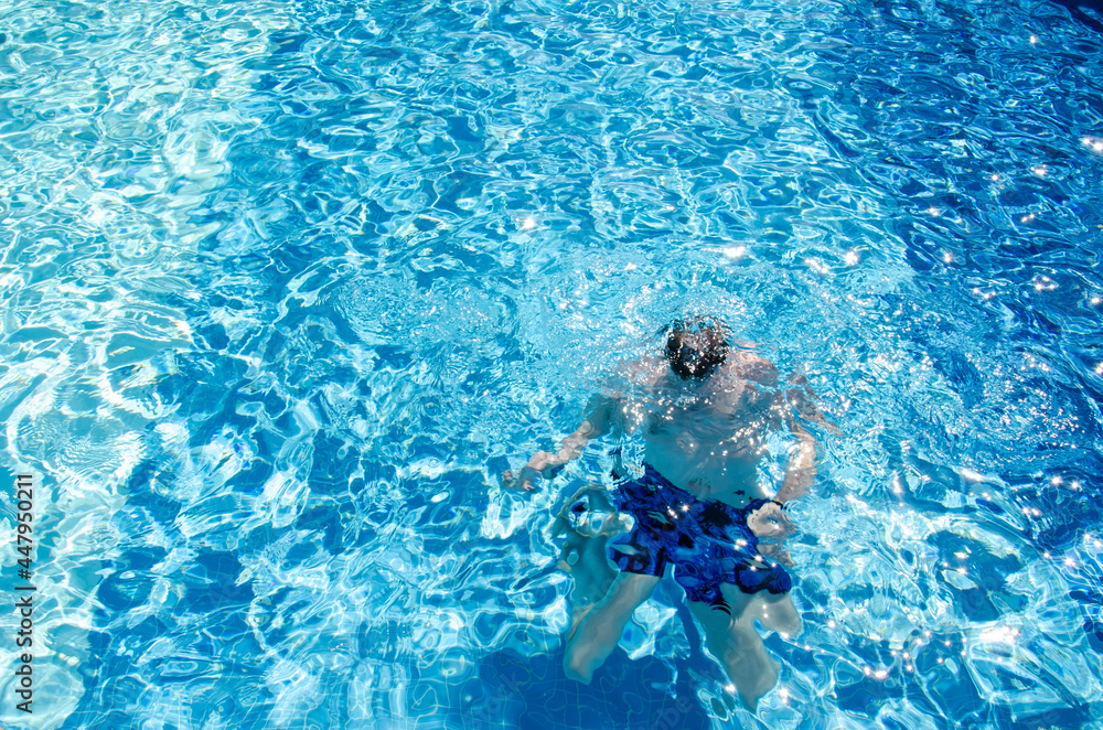 Caucasian man in swimming trunks under water dives in the pool with blue tiles on vacation. Adult male swims in swimming pool with azure water. Summer holidays concept.