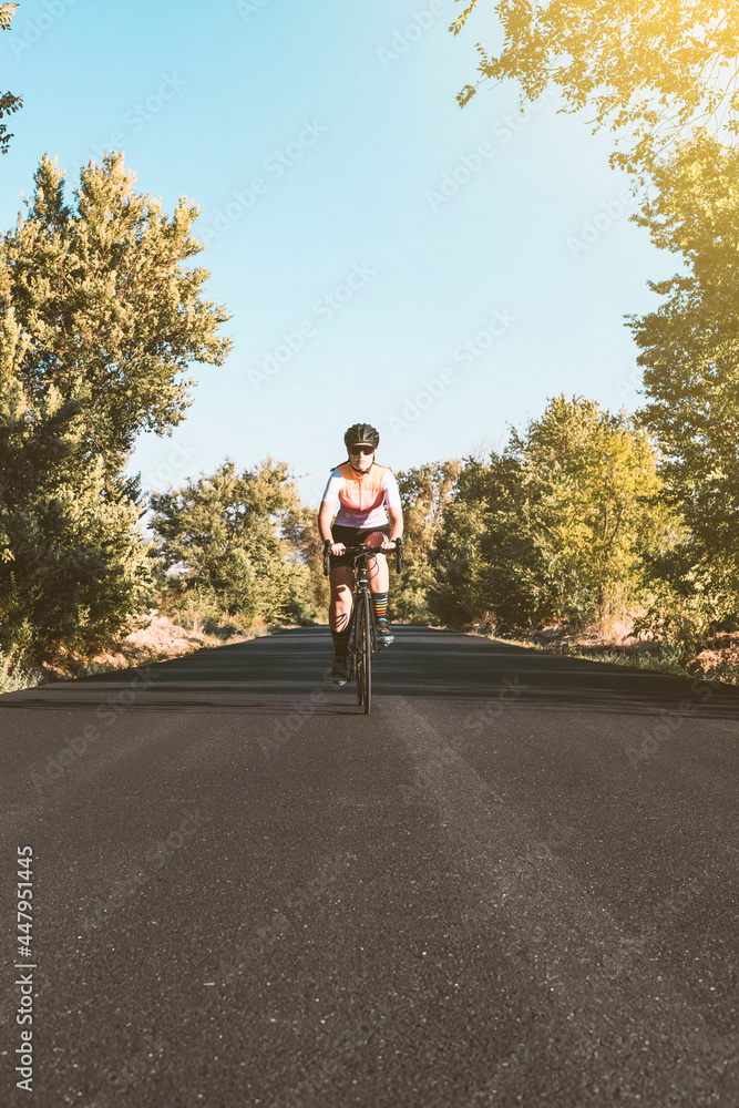 Fototapeta premium A young female cyclist riding her bike on the asphalt with trees on either side. Cycling concept. Sport concept.