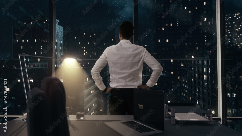 Back view of thoughtful Businessman standing in his office working late at night with hands on waist, looking out of the window as it rains contemplating his next big idea. Back view of thoughtful Businessman standing in his office working late at night with hands on waist, looking out of the window as it rains contemplating his next big idea.