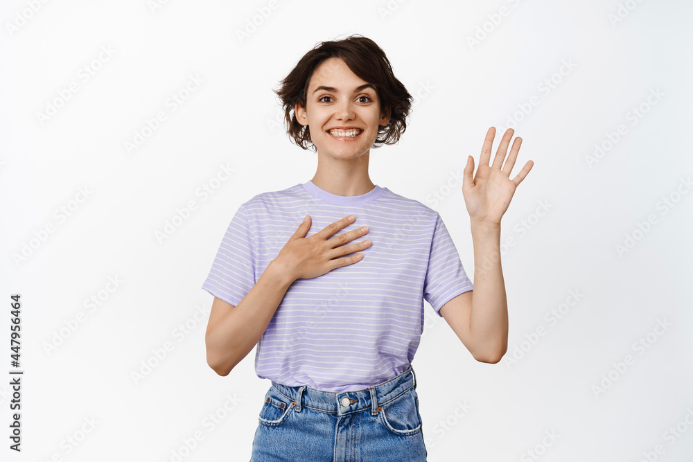 Friendly smiling woman hold arm on heart and palm raised high, making promise, introduce herself, my name is gesture, looking happy, standing over white background Friendly smiling woman hold arm on heart and palm raised high, making promise, introduce herself, my name is gesture, looking happy, standing over white background