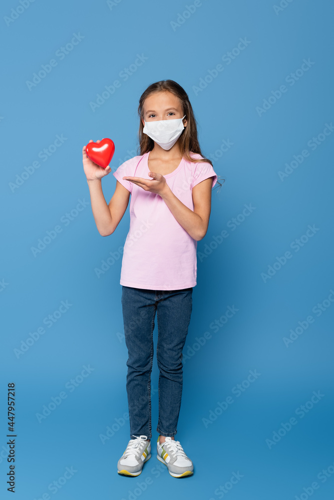 Girl in protective mask holding red decorative heart and pointing with hand on blue background Girl in protective mask holding red decorative heart and pointing with hand on blue background