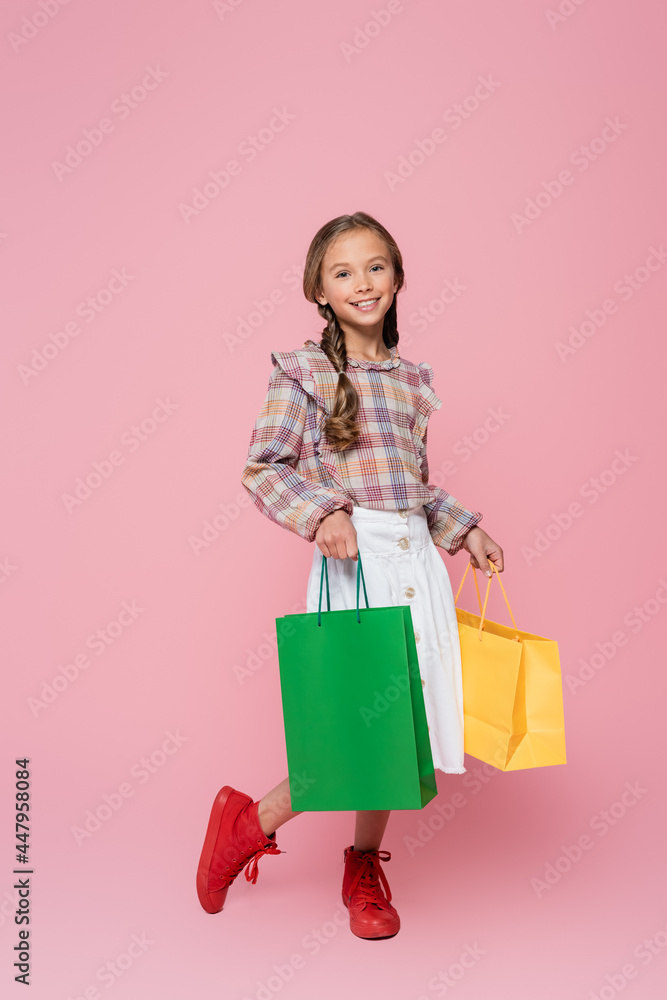 smiling child in stylish clothes holding green and yellow shopping bags on pink background