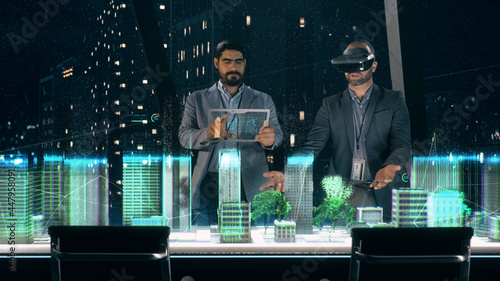 In the Near Future: Professional Designer in Suit wearing AR Headset presenting Architecture Project to Partner standing around Futuristic Table with Holographic Modern Augmented Reality Technology. photo
