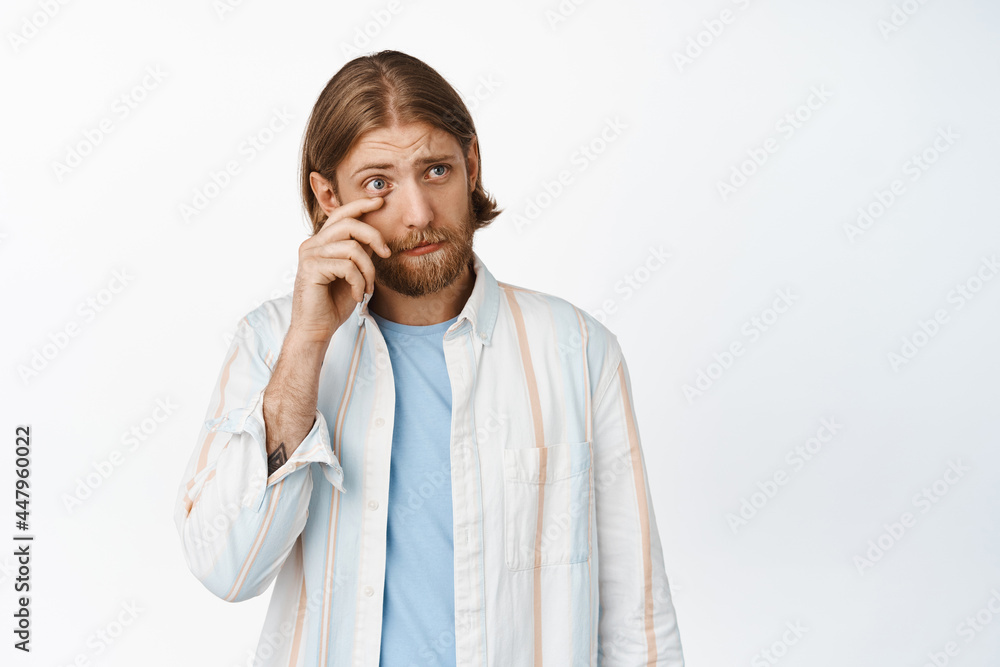 Young blond man sympathizing, crying, wiping tear below eye and looking pity aside, upset about something, feeling down depressed, standing against white background Young blond man sympathizing, crying, wiping tear below eye and looking pity aside, upset about something, feeling down depressed, standing against white background