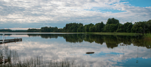 View of the lake with thick clouds in summer. A wooden footbridge enters the lake, and a reflection of clouds and trees can be seen in the water