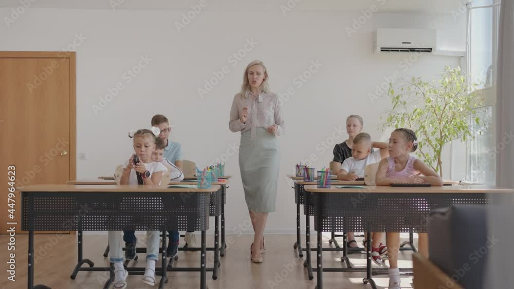 Female teacher controlling learning process in primary school. A female teacher walks between the desks and looks after the completion of the task Female teacher controlling learning process in primary school. A female teacher walks between the desks and looks after the completion of the task