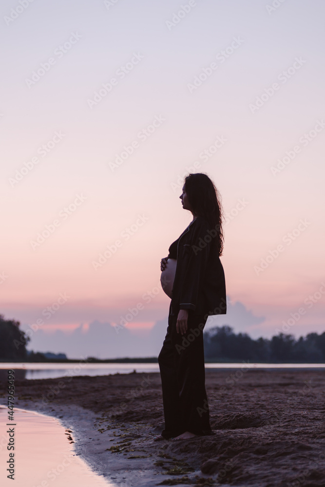 Silhouette of pregnant woman on beach. Beautiful young brunette with long curly hair stands on sand on beach and enjoys nature and setting sun. 