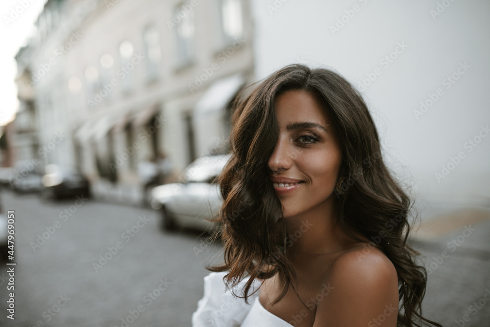 Portrait of charming young brunette with fluffy hair, tanned smooth skin and dark eyes, posing in city center, smiling and looking into camera Portrait of charming young brunette with fluffy hair, tanned smooth skin and dark eyes, posing in city center, smiling and looking into camera