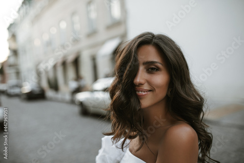 Portrait of charming young brunette with fluffy hair, tanned smooth skin and dark eyes, posing in city center, smiling and looking into camera