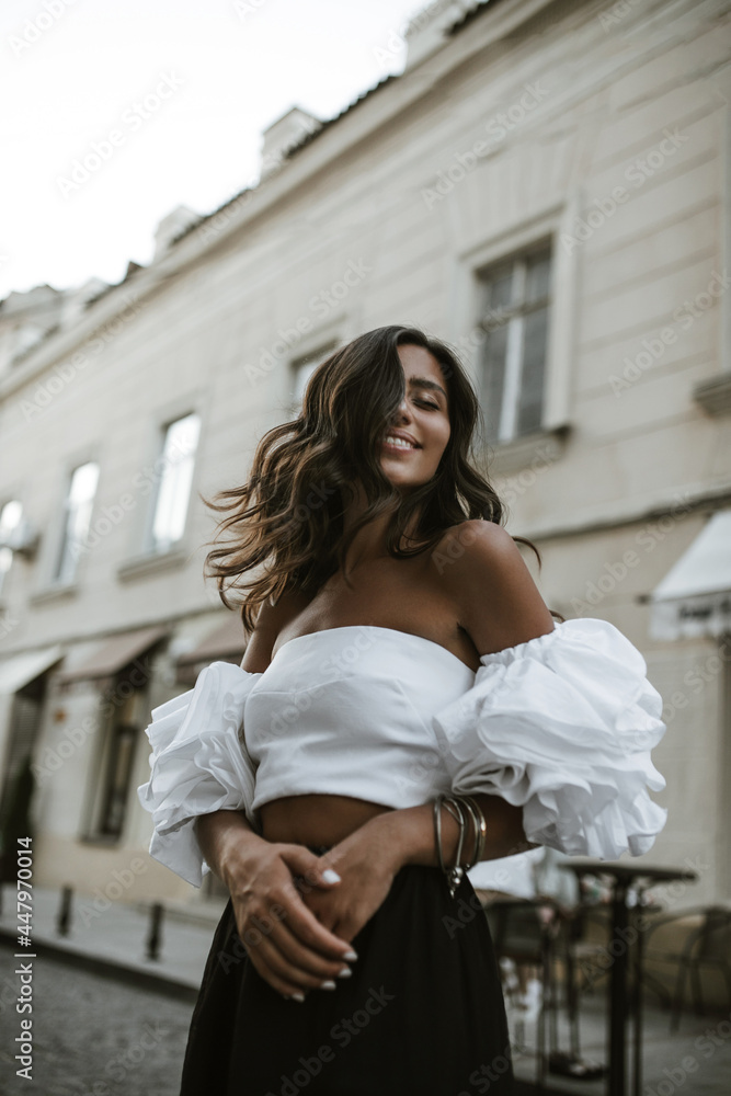 Cheerful young girl with brunette long hair in white off-shoulder blouse and dark pants smiling, waving head and walking in city