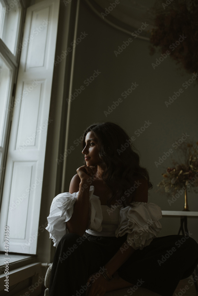 Portrait of charming young brunette in white off-shoulder top sitting in classic dark studio illuminated only from window with decorations against background Portrait of charming young brunette in white off-shoulder top sitting in classic dark studio illuminated only from window with decorations against background