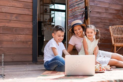 Mom or babysitter and two children, a girl and a boy, sitting at the door of a wooden house and watching cartoons on a laptop using wifi while relaxing outside the city