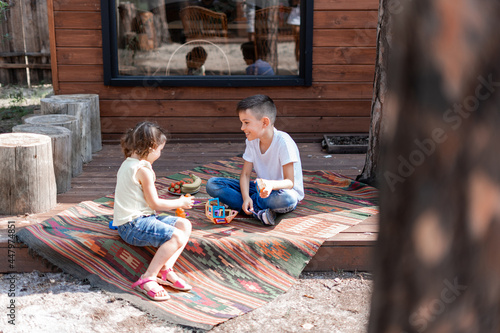 Little brother and sister sitting on the porch near a wooden house on an embroidered carpet, playing educational games, playing with a constructor, enjoying the summer photo