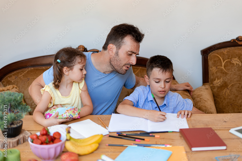 The father helps his son with school homework, makes sure that the boy writes correctly, while the youngest daughter sits in his arms.