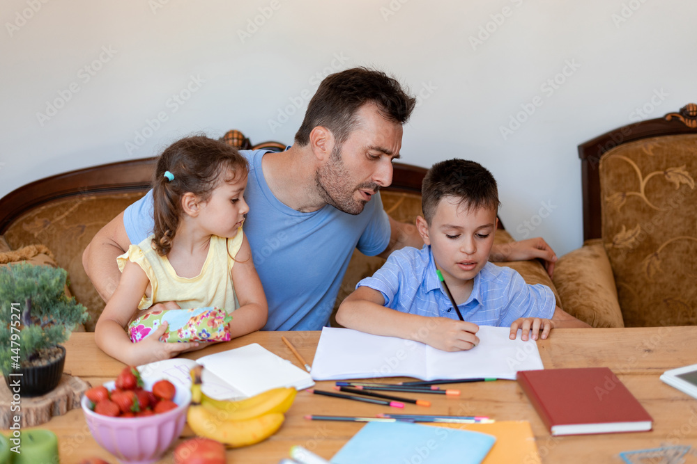 The father helps his son with school homework, makes sure that the boy writes correctly, while the youngest daughter sits in his arms. The father helps his son with school homework, makes sure that the boy writes correctly, while the youngest daughter sits in his arms.