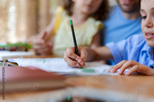 An elementary school student draws with pencils in a notebook or does school homework  under the close supervision of his sister and father.