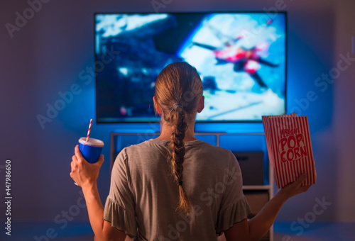 A young blonde woman in pajamas sits in front of a TV screen in the bedroom and watches her favorite movie. She is holding a pack of popcorn and a drink with a straw in a plastic glass. photo