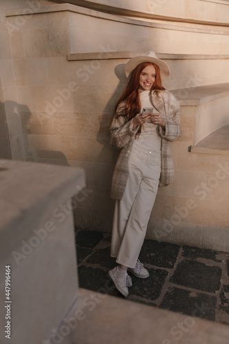 Full-length street photo of young girl holding mobile and standing against beige background outdoors. Gorgeous foxy lady wearing light clothes and stylish hat, smiling and looking straight forward