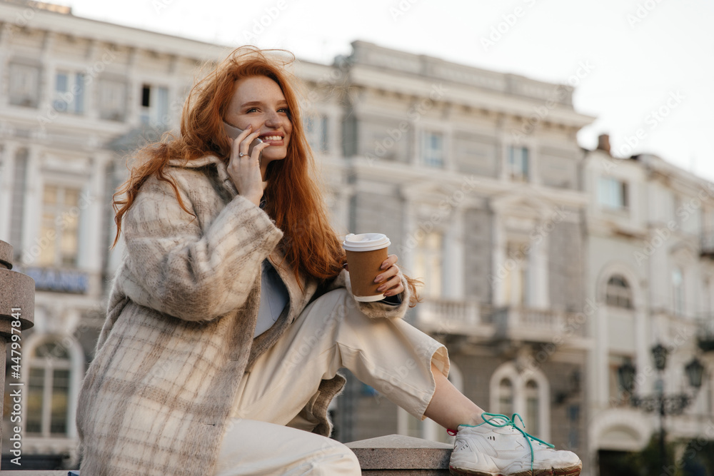 Close-up portrait of young foxy lady talking on phone in town. Adorable long-haired girl, wearing plaid coat and pants, posing against city landmark background