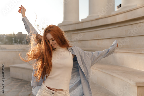 Cute foxy lady dancing outdoors at sunny day. Adorable young long-haired girl , wearing light turtleneck, blue shirt, moving and looking down against city landmark background