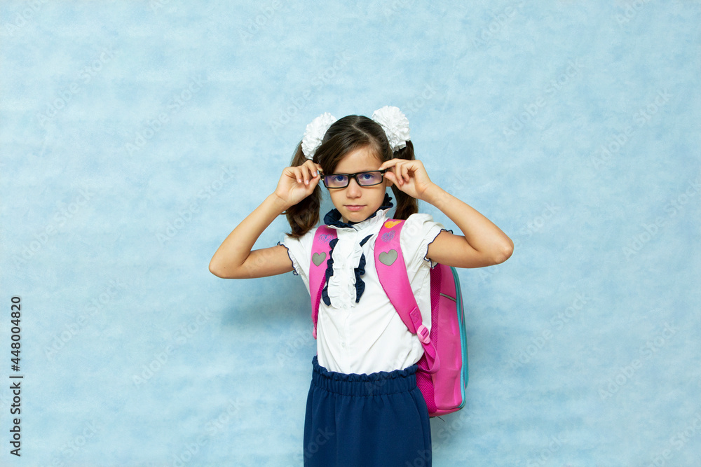 Funny dark-haired girl in glasses in school clothes with a backpack on her back on a blue background. Preparation for school. Place for your text. Funny dark-haired girl in glasses in school clothes with a backpack on her back on a blue background. Preparation for school. Place for your text.