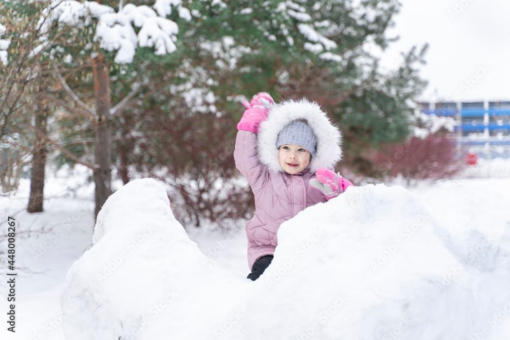 little girl in a warm coat and a hood plays snowballs in a snow fortress little girl in a warm coat and a hood plays snowballs in a snow fortress